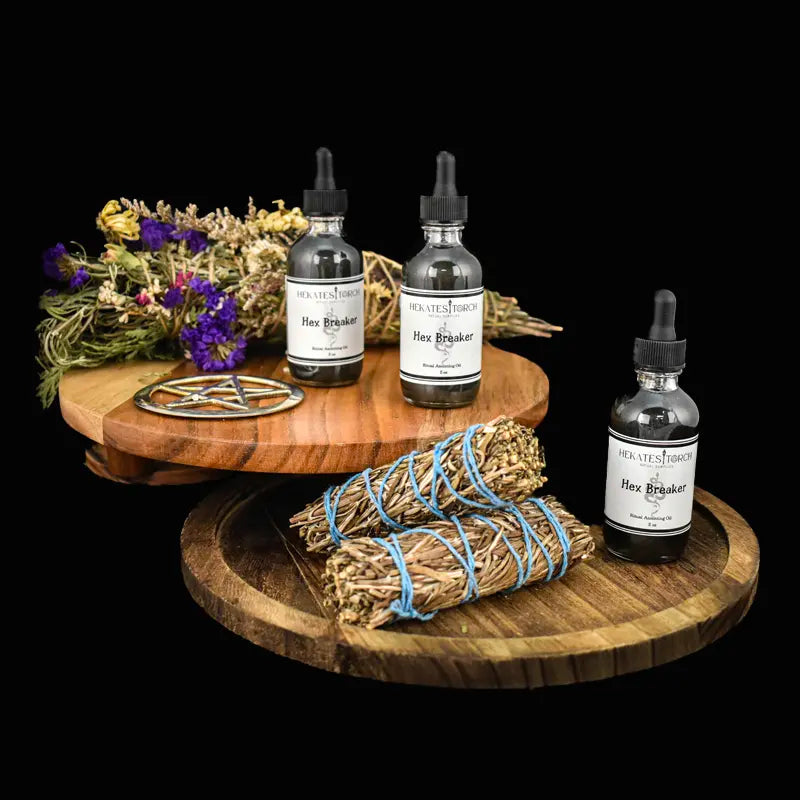 Three bottles of Hekates Hex Breaker Oil arranged on wooden trays with dried flowers and sage bundles against a black background